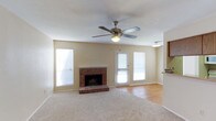 This single-floor home offers a comfortable and well-designed layout that balances functionality with classic charm. The living area serves as the heart of the home, anchored by a traditional brick fireplace with a raised brick hearth — a standout architectural feature that adds warmth and character to the room. Soft carpet flooring covers the main living area, transitioning to smooth hardwood-style flooring toward the adjacent spaces, creating a natural visual distinction between zones. A ceiling fan with a built-in light fixture is centrally mounted, promoting air circulation and providing ambient lighting throughout. Multiple windows dressed with blinds line the walls, inviting generous amounts of natural light while maintaining privacy. A glass-paneled door provides access to the exterior, further enhancing the sense of openness and connectivity to the outdoors. The neutral, light-toned walls offer a versatile backdrop that complements a wide range of interior styles. An adjoining area, visible from the living room, hints at additional functional spaces within the home. Overall, the space presents a cozy yet practical living environment with timeless details throughout.
