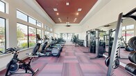 Located at 19815 Kenswick Drive in Humble, Texas, this fitness center is a thoughtfully designed single-floor facility that delivers a comprehensive workout environment. The space is defined by its architectural centerpiece: a recessed tray ceiling finished in a deep burgundy red, complemented by recessed lighting and a ceiling fan that together create a dynamic visual contrast against the crisp white surrounding walls. Large floor-to-ceiling windows line the perimeter, flooding the interior with abundant natural light and offering views of the surrounding outdoor amenity areas, including glimpses of a pool and lush greenery. The flooring throughout consists of multicolored modular carpet tiles in shades of red, pink, and gray, adding a modern, energetic aesthetic to the space. Full-length mirrors are mounted along one wall, enhancing the sense of openness and allowing users to monitor their form. The facility is generously outfitted with a wide array of equipment, including treadmills, elliptical machines, stationary bikes, and rowing machines arranged along the windowed wall, as well as a dedicated free-weight area stocked with a full dumbbell rack and weight benches. Cable machines and a squat/power rack station complete the strength training offerings. The overall ambiance is bright, spacious, and motivating.