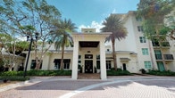This building presents a refined architectural design characterized by a crisp white exterior, standing metal roof panels, and a bold covered portico entrance supported by prominent square columns. The grand entryway creates an immediate sense of arrival, drawing visitors toward double glass doors framed by carefully curated landscaping. Tall, mature palm trees line the front of the property, complementing the tropical setting and reinforcing the building's upscale aesthetic. Neatly trimmed hedges and planted greenery border the façade, adding layers of texture and depth to the exterior presentation. Large windows with dark awning accents flank the entrance, allowing natural light to filter into the interior while contributing to the building's clean, symmetrical appearance. A paved brick forecourt extends from the entrance, providing a polished and well-defined approach to the building. The roofline features varied elevations, including a raised central section that adds visual interest and architectural dimension. The overall design conveys a professional, high-end character suited to its Florida setting, blending classical detailing with contemporary tropical influences in a cohesive and well-maintained presentation.