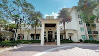 This building presents a refined architectural design characterized by a crisp white exterior, standing metal roof panels, and a bold covered portico entrance supported by prominent square columns. The grand entryway creates an immediate sense of arrival, drawing visitors toward double glass doors framed by carefully curated landscaping. Tall, mature palm trees line the front of the property, complementing the tropical setting and reinforcing the building's upscale aesthetic. Neatly trimmed hedges and planted greenery border the façade, adding layers of texture and depth to the exterior presentation. Large windows with dark awning accents flank the entrance, allowing natural light to filter into the interior while contributing to the building's clean, symmetrical appearance. A paved brick forecourt extends from the entrance, providing a polished and well-defined approach to the building. The roofline features varied elevations, including a raised central section that adds visual interest and architectural dimension. The overall design conveys a professional, high-end character suited to its Florida setting, blending classical detailing with contemporary tropical influences in a cohesive and well-maintained presentation.