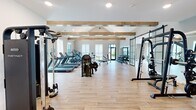 Located at 6170 Kitt Ln NW in Huntsville, Alabama, this fitness center offers a spacious, single-floor layout designed to accommodate a full range of workout activities. The space is defined by its striking vaulted ceiling accented with exposed wood beams, lending a warm yet modern aesthetic to an otherwise clean, neutral interior.

Light wood-tone flooring runs throughout, complementing the crisp white and soft gray walls. Large windows fitted with roller shades line the perimeter, flooding the space with natural light while offering views of the surrounding outdoor greenery. Black-framed glass barn doors serve as elegant dividers between zones, adding an industrial-chic design element.

The main workout floor is well-appointed with a full lineup of cardio equipment, including treadmills, ellipticals, and stationary bikes, alongside a robust selection of strength equipment such as a multi-station cable machine, Smith machine, weight bench, and a comprehensive dumbbell rack with large wall mirrors for form monitoring. A dedicated yoga and stretching room features oversized floor-to-ceiling windows, a ceiling fan, and open floor space. Additional amenities include a built-in water fountain station and convenient storage for equipment accessories, making this fitness center a thorough and thoughtfully designed facility.