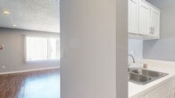 Located at 5345 N Sepulveda Blvd in Los Angeles, CA, this efficiently designed single-floor unit offers a thoughtful layout across its compact footprint. The living area is bright and open, featuring warm wood-look flooring that runs throughout the space, soft gray walls, and a textured ceiling with recessed lighting. A large window dressed with vertical blinds allows generous natural light to fill the room, complemented by a wall-mounted air conditioning unit for climate control. A ceiling fan is positioned near the kitchenette pass-through, which features white cabinetry and a countertop bar area, creating a functional division between the living and kitchen spaces. The bathroom is well-appointed, offering a long white vanity with ample cabinet storage, a wall-mounted mirror, and a separate shower stall with clean, bright finishes — all tied together by the same wood-look flooring found throughout the unit. The overall ambiance is clean, neutral, and move-in ready, with a cohesive color palette of whites and soft grays that lends a light, airy feel to every corner of the space.