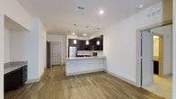 This single-floor space showcases a clean, modern design centered around an open-concept layout that seamlessly connects the kitchen and living area. The kitchen is a standout feature, equipped with dark espresso cabinetry that contrasts elegantly against the light-toned granite countertops. A peninsula-style breakfast bar extends the counter space and doubles as a casual dining or entertaining area. Stainless steel appliances, including a refrigerator, microwave, and range, add a contemporary, polished feel. Pendant lighting hangs above the peninsula, providing focused illumination and a stylish design accent, while recessed ceiling lights brighten the space evenly throughout. The flooring throughout is a warm, wood-look material that adds texture and a welcoming tone to the open space. Crisp white walls keep the atmosphere light and airy, enhancing the sense of openness. A utility entry door is visible near the kitchen, suggesting a practical layout with convenient access. Overall, the space presents a well-organized, functional design with modern finishes that create a comfortable and aesthetically cohesive living environment.