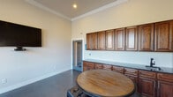 This single-floor communal space is designed as a comfortable and functional lounge or gathering room, combining practical amenities with an inviting atmosphere. The room is defined by its impressively tall ceilings accented with crown molding, giving the space a sense of openness and grandeur. Polished concrete flooring runs throughout, lending a modern, durable finish that complements the warm tones of the rich, dark wood cabinetry lining one wall.

The built-in cabinetry features both upper and lower cabinets with detailed raised-panel doors, paired with a dark stone countertop and an undermount sink — forming a well-equipped kitchenette ideal for serving refreshments. Multiple round wooden tables surrounded by black leather-upholstered stools are arranged throughout the room, providing ample casual seating for groups.

Large multi-pane windows draped with neutral curtains allow generous natural light to fill the space while offering views of the surrounding greenery and outdoor areas. A wall-mounted flat-screen television serves as a focal point on one end of the room, enhancing its use as an entertainment or meeting space. A piece of decorative artwork adds a touch of character near the exit door. The space is complete with a direct exterior door, providing convenient access to the outdoors.