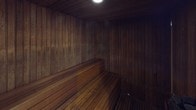 This electric dry sauna is a dedicated heat therapy space constructed entirely with traditional wood craftsmanship. Every surface — the walls, ceiling, and floor — is clad in vertical wood planking that has developed a deep, rich patina over time, contributing to the space's warm and enveloping character. The interior is compact and efficiently designed, with tiered wooden benches arranged along the walls to accommodate multiple users at varying heat levels, as warm air naturally rises toward the upper benches. A single overhead light provides soft, subdued illumination, reinforcing the calm and restorative ambiance of the space. The sauna operates as a dry sauna, powered by an electric heating unit fitted with decorative rocks, and signage clearly advises against pouring water on the rocks. Entry is through a full-height door that separates the sauna from an adjacent wet area. The consistent use of natural wood throughout creates a seamless, immersive environment that is both functional and aesthetically cohesive, offering a traditional sauna experience in a well-appointed setting.