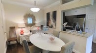 This well-appointed single-floor residence exudes a refined yet welcoming atmosphere, thoughtfully designed with a cohesive aesthetic throughout. The dining area serves as a central highlight, anchored by a sleek oval white dining table surrounded by upholstered grey chairs that offer both comfort and sophistication. A pendant light fixture hangs overhead, casting a warm glow over the space and drawing attention to the room's crown molding details that frame the ceiling with classic elegance. A striking sunburst mirror adorns one wall, complementing a bold abstract canvas painting that adds a dynamic focal point to the room. Decorative glass bowls and floral accents on the tabletop contribute to the curated, inviting feel of the space. A coordinating sideboard with lamps and decorative objects adds further depth and function to the dining area. Hardwood flooring runs throughout, grounding the space with warmth and continuity. A pass-through opening connects the dining area to the kitchen, which features dark cabinetry, light stone countertops, and stainless steel fixtures — offering both visual contrast and practical accessibility between the two spaces. The overall ambiance is polished and tastefully styled.
