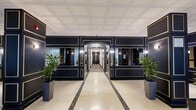 Located at 3435 Rue Drummond in the heart of Montréal, this sophisticated lobby makes a striking first impression with its refined, classical design. The space is finished with large-format cream marble flooring accented by inlaid dark marble border strips, lending a sense of grandeur and polish throughout.

Commanding navy-blue paneled walls trimmed in gold define the interior architecture, incorporating mirrored insets that reflect light and amplify the sense of openness. Tall, boxy structural columns dressed in the same dark paneling with gold detailing serve as bold focal points within the space.

Natural light filters generously through oversized glass entry doors and floor-to-ceiling windows dressed with layered sheer and patterned drapes, creating a bright, welcoming atmosphere. A recessed tile ceiling with integrated pot lighting complements the overall illumination.

The lobby is thoughtfully furnished with tall dark planters holding lush tropical greenery, adding life and warmth to the formal setting. A classical sculptural bust displayed on a white pedestal contributes an artistic touch. A dedicated mailbox area with polished metal units is neatly integrated along one wall, ensuring practical functionality without compromising the aesthetic. The overall ambiance is one of understated luxury and timeless elegance, befitting a distinguished Montréal address.
