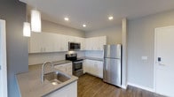 This single-floor space showcases a contemporary kitchen designed with both functionality and style in mind. The kitchen features a clean, modern aesthetic highlighted by crisp white cabinetry that extends from floor to ceiling, providing generous storage throughout. Stainless steel appliances, including a refrigerator, electric range, and over-the-range microwave, lend a polished, cohesive look to the space. The countertops are finished in a neutral gray tone, complementing the overall color palette. A stainless steel double-basin sink is positioned at the peninsula, which also serves as a natural divider between the kitchen and the adjoining area, offering additional workspace and casual seating potential. Recessed lighting is evenly distributed across the ceiling, ensuring the space is well-lit throughout. The flooring features warm-toned hardwood-style planks that contrast tastefully with the light cabinetry and walls. A small window allows for a touch of natural light to enter. The overall ambiance is clean, bright, and functional, reflecting a thoughtfully designed modern living space.