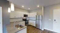 This single-floor space showcases a contemporary kitchen designed with both functionality and style in mind. The kitchen features a clean, modern aesthetic highlighted by crisp white cabinetry that extends from floor to ceiling, providing generous storage throughout. Stainless steel appliances, including a refrigerator, electric range, and over-the-range microwave, lend a polished, cohesive look to the space. The countertops are finished in a neutral gray tone, complementing the overall color palette. A stainless steel double-basin sink is positioned at the peninsula, which also serves as a natural divider between the kitchen and the adjoining area, offering additional workspace and casual seating potential. Recessed lighting is evenly distributed across the ceiling, ensuring the space is well-lit throughout. The flooring features warm-toned hardwood-style planks that contrast tastefully with the light cabinetry and walls. A small window allows for a touch of natural light to enter. The overall ambiance is clean, bright, and functional, reflecting a thoughtfully designed modern living space.