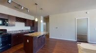 Located at 1200 E Parmer Lane in Austin, Texas, this single-floor residence offers a well-designed, open layout that seamlessly connects the kitchen and living areas. The kitchen is a functional centerpiece, outfitted with rich dark wood cabinetry, granite-style countertops, a full suite of black appliances including a range, microwave, refrigerator, and dishwasher, as well as a built-in wine rack and a peninsula island with pendant lighting above. Track lighting overhead adds to the kitchen's practicality. Hardwood-style flooring flows throughout the main living and kitchen areas, while a low half-wall divider elegantly defines the living space. The living room benefits from natural light provided by a large window, complemented by a ceiling fan for year-round comfort. The bedroom features plush carpet flooring, generous natural light from a wide window bank, and its own ceiling fan. The hallway connects to the bathroom, which is finished with a dark wood vanity, granite-style countertop, and a tiled shower/tub combination. A dedicated electrical panel is accessible from the hallway. The overall design palette—neutral walls, dark cabinetry, and warm wood-tone floors—creates a cohesive and inviting atmosphere throughout.