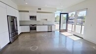 This contemporary 3-bedroom, 4-bathroom townhome at 2496 S Centinela Ave, Unit 1, Los Angeles, CA 90064, offers 1,310 square feet of modern living across three floors. The ground floor entry level (190 sq ft) features polished concrete flooring and a sculptural open-riser staircase with glass panel railings, connecting all three levels. The main living floor (550 sq ft) showcases an open-plan kitchen and living area with polished concrete floors, sleek white cabinetry, stainless steel appliances, and recessed lighting throughout. The upper floor (570 sq ft) houses the bedrooms, including a 170 sq ft primary suite with wide-plank hardwood flooring, a private balcony accessible through floor-to-ceiling glass doors offering neighborhood views, and an en-suite bathroom featuring a glass-enclosed tile shower. Additional bathrooms include a full bath with tub and vessel sink. A standout feature is the expansive rooftop deck with a colorful paneled railing, open-sky views of the surrounding Los Angeles neighborhood, and palm tree vistas—ideal for outdoor entertaining. Natural light flows throughout the home via large windows and glass doors.