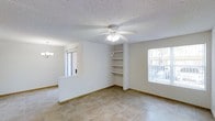 This single-story space offers an open and functional layout filled with natural light from multiple windows. The main living area features neutral tile flooring throughout, white walls, and a textured ceiling, contributing to a clean and understated aesthetic. A ceiling fan with an integrated light fixture is centrally positioned, providing both ventilation and ambient lighting. Built-in shelving along one wall offers convenient storage and display space, adding a practical architectural detail to the room. A partial wall divides the living area from an adjacent space, maintaining an open feel while subtly defining separate zones. A chandelier is visible in the neighboring area, hinting at a dedicated dining or entryway space. Large windows on multiple walls allow ample natural light to filter in, enhancing the bright and open atmosphere. The overall design is straightforward and move-in ready, with neutral finishes that provide a versatile backdrop suited to a variety of interior styles.