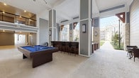 This expansive community lounge is designed to seamlessly blend indoor comfort with outdoor living. The space features soaring ceilings and an open-air layout framed by striking limestone and gray shiplap accent walls, creating a sophisticated yet relaxed atmosphere throughout. String lights draped overhead add a warm ambiance, while large ceiling fans provide comfort across the generous gathering areas. A centerpiece billiards table with a bold blue felt surface anchors the main entertainment zone, complemented by a dedicated bar counter with dark wood cabinetry and cushioned high-back bar seating. An outdoor kitchen area showcases marble-topped counters with a deep sink, ideal for hosting and entertaining. A built-in lounge nook features a continuous wraparound bench upholstered in rich blue cushions, offering a comfortable spot to relax while taking in courtyard views through the open-air columns. A limestone fireplace serves as a dramatic focal point, topped with a wall-mounted flat-screen television — perfect for communal gatherings. Light-toned tile flooring runs throughout, tying the various zones together with a clean, modern aesthetic. The lounge opens directly to a landscaped courtyard, with views of the surrounding residential buildings completing the inviting, resort-style setting.