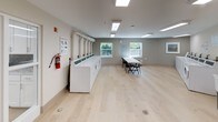 Located at 500 Harry S Truman Dr in Largo, Maryland, this shared laundry facility is a well-organized, single-floor space designed for practical, communal use. The room features a generous open layout with light hardwood-style flooring throughout, contributing to a clean and welcoming atmosphere.

Along one wall, a row of top-loading washing machines is neatly aligned, accompanied by posted instructional guides to assist users. On the opposite side, a matching row of front-loading dryers is equipped with ventilation piping routed along the wall, ensuring proper airflow and functionality. A centrally placed folding table with chairs provides a convenient surface for sorting and folding laundry.

The space is illuminated by recessed fluorescent panel lighting in the ceiling, ensuring bright, even coverage throughout. Windows along one wall allow natural light to filter in, offering views of the surrounding greenery and adding to the room's airy feel. Additional amenities include a payment kiosk station, a garment hanging rack, a paper towel dispenser, and a waste bin for user convenience. A fire extinguisher is mounted on the wall in compliance with safety standards. The neutral, light-toned walls and uncluttered design make this facility easy to navigate and maintain.