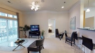 This single-floor residence offers a thoughtfully designed open-concept layout that seamlessly blends the living and dining areas into one cohesive, welcoming space. The living room is anchored by plush wall-to-wall carpeting in a warm neutral tone, complemented by crisp white walls that create a bright and airy ambiance. A ceiling fan with integrated lighting fixture provides both comfort and illumination, while recessed lighting adds a polished finishing touch overhead. The space is furnished with sleek leather seating and a glass-top coffee table, lending a contemporary feel to the room. A flat-screen television on a dark media console serves as the focal point of the entertainment area, while a decorative potted tree adds a touch of natural greenery. Adjacent to the living area, a compact dining nook features a round glass-top table paired with dark upholstered chairs, ideal for intimate meals. A soft, abstract wall art piece enhances the dining corner with subtle color. An open doorway leads seamlessly into an additional room, offering a sense of depth and connectivity throughout the floor plan. Overall, the space exudes a clean, modern aesthetic with a calm and comfortable atmosphere.