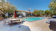 This two-story residential complex is designed around a resort-style outdoor amenity area that serves as its central feature. The architecture blends a Mediterranean-influenced clubhouse — distinguished by a classic terracotta tile roof and arched openings — with a more contemporary multi-story residential building that offers private balconies and exterior staircase access between floors.

At the heart of the outdoor space is a generously sized swimming pool with clear, blue-green water, framed by brick edging and a wide concrete sun deck. The deck is well-appointed with multiple rows of lounge chairs and chaise lounges on both sides of the pool, along with umbrella-shaded tables and benches that provide covered seating options. A cozy pod-style lounging chair adds a distinctive design touch near the pool's edge.

The outdoor area is enclosed by an iron security fence with a gated entrance, accented by a decorative lantern-style lamp post and surrounded by mature deciduous trees that provide natural shade and seasonal greenery. Views beyond the fence reveal rolling hills and open natural landscape, lending a serene, retreat-like atmosphere to the property. The combination of functional amenities, thoughtful landscaping, and scenic surroundings makes this outdoor space a standout feature of the complex.