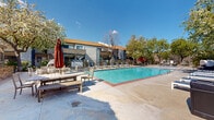 This two-story residential complex is designed around a resort-style outdoor amenity area that serves as its central feature. The architecture blends a Mediterranean-influenced clubhouse — distinguished by a classic terracotta tile roof and arched openings — with a more contemporary multi-story residential building that offers private balconies and exterior staircase access between floors.

At the heart of the outdoor space is a generously sized swimming pool with clear, blue-green water, framed by brick edging and a wide concrete sun deck. The deck is well-appointed with multiple rows of lounge chairs and chaise lounges on both sides of the pool, along with umbrella-shaded tables and benches that provide covered seating options. A cozy pod-style lounging chair adds a distinctive design touch near the pool's edge.

The outdoor area is enclosed by an iron security fence with a gated entrance, accented by a decorative lantern-style lamp post and surrounded by mature deciduous trees that provide natural shade and seasonal greenery. Views beyond the fence reveal rolling hills and open natural landscape, lending a serene, retreat-like atmosphere to the property. The combination of functional amenities, thoughtful landscaping, and scenic surroundings makes this outdoor space a standout feature of the complex.