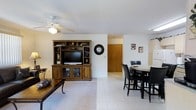 This single-floor residence offers a comfortable and practical layout centered around an open-concept living and dining area. The living space is anchored by a large wood entertainment center with built-in shelving, cabinetry, and display niches, providing ample storage and a focal point for the room. A ceiling fan with integrated lighting ensures year-round comfort and adds a classic touch to the space.

The flooring transitions seamlessly between plush carpeting in the living area and light-toned ceramic tiles in the dining and adjoining spaces, creating a clear yet open delineation between zones. The dining area accommodates a round table with seating for four, complemented by dark-framed chairs that contrast nicely with the neutral walls and bright surroundings.

The interior is bathed in soft, even lighting, enhanced by the crisp white walls and ceilings that reflect light throughout the space. A sliding closet door is visible in the background, hinting at additional storage options nearby. The overall ambiance is clean, bright, and functional, making it well-suited for everyday living. The open sightlines between rooms contribute to a spacious feel, while the warm wood tones of the furnishings add a welcoming and homey character throughout.