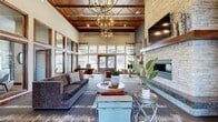 This single-floor space offers a thoughtfully designed open-concept layout that seamlessly blends a living area, kitchen, and entrance into one cohesive environment. The living room immediately commands attention with its soaring ceilings, featuring a striking wood-paneled ceiling section accented by an oversized, sculptural orb chandelier that serves as a focal point of the room. A floor-to-ceiling stacked stone fireplace anchors one wall, complete with a built-in television above a rustic wood mantel, creating a warm and inviting atmosphere. Large, multi-pane windows line the perimeter, flooding the space with abundant natural light and offering views of the surrounding exterior. The entrance features double French doors framed by transom windows, flanked by contemporary decorative wall accents and lush tropical plants, setting an welcoming tone. The kitchen is well-appointed with warm wood cabinetry, white countertops, stainless steel appliances, and a detailed mosaic tile backsplash, complemented by light hardwood flooring. The dining area, positioned near the kitchen, features a marble-topped round table with modern leather chairs beneath pendant lighting. Throughout, the design balances rustic elements — exposed wood, natural stone — with modern furnishings, creating a refined yet comfortable communal atmosphere.