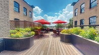 Located at 330 E Ninth St in Saint Paul, Minnesota, this generously sized rooftop deck offers an impressive outdoor retreat that combines classic urban architecture with welcoming open-air amenities. The deck is constructed with warm-toned hardwood planking that extends across a substantial footprint, creating an inviting environment for gathering and relaxation.

The space is thoughtfully arranged with multiple seating areas, each featuring wrought iron and cushioned patio furniture grouped around circular tables and shaded by bold red umbrellas. These clusters are distributed to encourage both intimate conversation and larger social gatherings. Raised planter beds line the perimeter, filled with colorful seasonal flowers and lush greenery that add a natural vibrancy to the setting.

The deck is flanked on both sides by the original multi-story brick masonry of the surrounding building, with large multi-pane windows lending a historic industrial character to the space. A sleek cable railing system defines the outer edge, allowing unobstructed views of the Saint Paul skyline and surrounding neighborhood. Grilling stations positioned near the building entrance add practical functionality. The open sky above ensures abundant natural light throughout the day, making this rooftop deck a standout communal feature of the property.