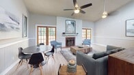 This single-floor space is designed with an open, airy layout that combines a living area, dining nook, and kitchen in a cohesive, functional arrangement. The living area serves as the focal point, showcasing soaring vaulted ceilings that amplify the sense of space and volume. A wood-burning fireplace with a classic white mantel anchors one wall, flanked on either side by wood-framed French doors that invite natural light and provide views of the surrounding greenery. A ceiling fan overhead adds both practicality and a relaxed ambiance to the room.

Light wood-style flooring runs throughout, lending warmth and visual continuity across the open floor plan. The dining area features a round table with modern chairs, positioned to take advantage of the natural light streaming through the French doors. A console table behind the seating area offers additional functionality without disrupting the flow of the space.

The kitchen is defined by a peninsula breakfast bar with seating for three, creating a natural transition between the cooking and living areas. Gray cabinetry, a decorative tile backsplash, and black appliances give the kitchen a clean, contemporary character. A dedicated mechanical room is conveniently tucked nearby. Overall, the space presents a welcoming, well-organized environment with thoughtful design details throughout.