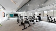 Located at 1900 SW 13th St in Gainesville, Florida, this modern fitness center offers a thoughtfully designed exercise space across a single floor. The layout is generously proportioned, providing ample room for a diverse range of workout activities. The interior is defined by a striking two-tone ceiling design — a textured dark gray central section contrasted against crisp white surrounding areas — creating a dynamic architectural focal point overhead. Sleek linear pendant lighting complements the ceiling fans distributed throughout, ensuring the space remains bright and well-ventilated. Patterned, light-toned carpet tiles cover the flooring throughout, adding texture and comfort underfoot. The exercise area is outfitted with a fleet of Spinning-brand cycling bikes, elliptical machines, and various cardio equipment, along with a dumbbell and kettlebell rack for strength training. A wall-mounted TRX suspension training system offers additional versatility. A full floor-to-ceiling mirror wall anchors one end of the room, paired with a mounted flat-screen television and motivational athletic artwork on the white walls. Large windows along the perimeter invite natural light and views of the surrounding outdoors. A dedicated open studio section with yoga mats provides space for stretching and floor-based workouts, making this fitness center a comprehensive facility for varied training needs.