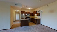 Located in Ankeny, Iowa, this single-story space showcases a thoughtfully designed open-concept layout that seamlessly integrates the kitchen and living areas. The kitchen serves as the focal point of the space, featuring rich, dark wood cabinetry that extends to the upper walls, offering generous storage throughout. A central island provides additional workspace and a natural gathering point, illuminated by sleek pendant lights hanging at varying heights. The kitchen is equipped with a full suite of stainless steel appliances, including a refrigerator, electric range, built-in microwave, and dishwasher, alongside a countertop sink positioned along the perimeter cabinetry. An elegant chandelier with multiple globe lights adds a refined touch above the dining area. The adjoining living space is finished with neutral carpet, creating a warm and comfortable atmosphere that complements the kitchen's darker tones. A hallway visible from the main living area leads to additional rooms, suggesting a well-organized floor plan. Neutral wall colors and consistent flooring choices throughout contribute to a cohesive and inviting interior.