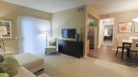 This single-floor residence offers a thoughtfully designed layout that balances comfort and functionality. The living area is spacious and inviting, featuring plush wall-to-wall carpeting, soft neutral wall tones, and large vertical blinds that allow for generous natural light. A dedicated media wall with ample storage anchors the room, providing a natural focal point for relaxed gatherings or quiet evenings at home. The space transitions smoothly into the kitchen and hallway corridor, where warm hardwood-style flooring takes over, offering a clean contrast to the carpeted living zone. The kitchen is equipped with a stainless steel refrigerator and benefits from efficient overhead lighting, contributing to a bright and practical cooking environment. A hallway connects the main living spaces to the private areas of the home, maintaining a clear and intuitive flow throughout. The overall ambiance is calm and understated, with a neutral color palette, clean lines, and consistent finishes that lend the space a cohesive, move-in-ready feel. The combination of soft textures, practical storage, and an open sightline from the living area into the adjacent spaces makes this residence both comfortable and highly functional for everyday living.