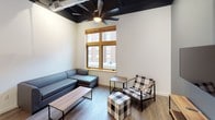 This single-floor unit blends industrial and contemporary design elements throughout its thoughtfully arranged layout. The living area serves as the visual centerpiece, boasting soaring ceilings with exposed ductwork and dark-painted structural elements that lend an urban loft character. Large wood-framed windows flood the space with natural light and offer views of the surrounding cityscape. A ceiling fan with a built-in light fixture anchors the room, which is furnished with a sectional sofa, plaid accent chair with ottoman, and a wall-mounted television on a wood-and-metal media console.

The kitchen features white shaker cabinetry, a classic subway tile backsplash, and a generous white countertop peninsula with bar-height seating for casual dining. Stainless steel appliances and matte black pendant lighting complete the modern aesthetic, while the exposed ceiling above maintains the industrial loft feel throughout the common areas.

Multiple bedrooms each feature dark hardwood-style flooring, ceiling fans, and neutral white walls, with select rooms benefiting from natural light through wood-framed windows. The bathroom is finished in crisp white with wood-look flooring, a white vanity, and matte black hardware and towel bars, creating a clean and cohesive aesthetic.