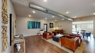 This leasing office and meeting room in Baton Rouge, Louisiana, is a thoughtfully designed single-floor space that balances professional functionality with a welcoming, contemporary aesthetic. Rich hardwood flooring extends throughout the main areas, anchoring the warm and inviting atmosphere.

The primary lounge area serves as the central hub of the space, furnished with generously sized cognac leather sofas and club chairs arranged around a substantial industrial-style coffee table atop a woven area rug. Curated artwork, including bold colorful canvases and decorative wall sculptures, animates the neutral walls. An exposed metallic HVAC duct runs across the ceiling, lending an industrial design element, while recessed lighting ensures the space is well-illuminated.

A dedicated workstation area with a desk and seating provides space for leasing staff. Adjacent to the main lounge, a separate meeting room accommodates a large wood dining-style conference table surrounded by upholstered chairs, with a vibrant abstract painting as a focal point and natural light filtering through a window.

A compact kitchenette features grey cabinetry, a white countertop island, stainless steel appliances, and light wood flooring. A small casual seating area with a round table and teal chairs completes this section. Natural light is accessible from multiple points, and a glazed door connects to an adjacent fitness area.