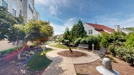 Located in Alexandria, Virginia, this outdoor dog park offers a thoughtfully designed green space for pets and their owners. The park features a winding concrete pathway that curves through lush, well-maintained lawns and carefully landscaped mulched garden beds filled with a variety of shrubs, ornamental grasses, and mature trees that provide ample shade. A central circular turf area serves as an open play zone, anchored by a dog agility sculpture that adds a charming, functional focal point to the space. Comfortable seating benches are positioned along the pathway, offering resting spots for owners while their pets explore. The perimeter is defined by a combination of wooden privacy fencing and black metal fencing, creating a secure and enclosed environment. Pet waste stations equipped with bag dispensers and waste bins are conveniently located throughout the park, promoting cleanliness and responsible pet ownership. The surrounding multi-story residential buildings in brick and neutral siding frame the space, providing a sense of community integration. Mature evergreen and deciduous trees line the boundaries, offering natural screening and a serene, park-like atmosphere. The open sky above allows for abundant natural light, making this a bright and welcoming outdoor amenity.