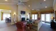 This single-floor space exudes a classic, welcoming charm defined by its striking architectural details and thoughtfully designed living area. The great room immediately draws the eye upward to its soaring vaulted ceilings, adorned with crown molding and equipped with dual ceiling fans that enhance both comfort and visual appeal. Recessed lighting complements the warm, golden-yellow walls, creating a bright and cozy atmosphere throughout.

A traditional white-mantled fireplace serves as the room's central focal point, framed by a brick hearth and topped with a wall-mounted flat-screen television — a seamless blend of classic and contemporary design. Elegant wall sconces flank the fireplace, adding a refined, ambient glow to the space.

Large French doors along the exterior wall flood the room with generous natural light and offer views to the outdoors, while fabric valances above each window provide a polished finishing touch. The living area is generously proportioned, comfortably accommodating a plush sofa, upholstered armchairs, and a round wooden coffee table arranged atop a decorative area rug. The overall ambiance is warm, traditional, and inviting — ideal for both relaxed everyday living and entertaining guests.