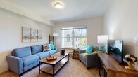 This single-floor residence offers a thoughtfully organized layout encompassing a living area, kitchen, two bedrooms, and a full bathroom. The living room serves as the central gathering space, finished with plush carpeting and a large window that invites natural light and provides views of the surrounding exterior. The bright, neutral walls and open feel create a welcoming ambiance throughout.

The kitchen is designed for practical use, arranged in an L-shaped configuration with light wood laminate flooring, cream cabinetry providing ample storage, and white countertops. Black appliances — including an electric range, dishwasher, and refrigerator — offer a striking contrast against the lighter finishes. A built-in range hood and a stainless steel sink complete the workspace.

Both bedrooms are carpeted and comfortably sized to accommodate full or queen beds. Each room features generous closet space with bi-fold doors, and windows allow for natural light. The neutral tones throughout maintain a calm, restful atmosphere.

The bathroom is elongated and efficient, featuring warm wood-tone laminate flooring, a wide vanity with ample drawer storage, a large mirror, and a shower-tub combination. Overall, the unit presents a clean, functional design with a cohesive, understated aesthetic.