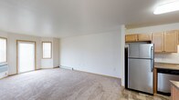 This single-floor space presents an open and functional layout designed for comfortable everyday living. The main living area is finished with wall-to-wall carpeting in a neutral tone, contributing to a clean and understated aesthetic throughout. White walls and a smooth white ceiling help maximize the sense of openness, while a window along the far wall allows natural light to filter into the room, brightening the interior. A baseboard heater runs along the base of the main wall, providing an efficient heating solution integrated discreetly into the space.

The kitchen is positioned adjacent to the living area and transitions from carpet to tile flooring, visually distinguishing the two zones. Light wood cabinetry offers ample storage overhead, and a stainless steel refrigerator anchors the kitchen, lending a modern touch to the overall design. The open sightlines between the kitchen and living space create a connected feel, making the layout practical for daily use.

Overall, the space maintains a straightforward, move-in-ready character with neutral finishes throughout, offering a versatile backdrop well-suited to a variety of furnishing and décor styles.