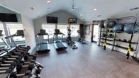 This fitness center offers a clean, modern environment designed to support a full range of workout routines. The space is defined by its vaulted ceiling, which lends an open, airy feel, while recessed lighting and a ceiling fan ensure comfort during exercise. Large windows line the walls, flooding the room with natural light and providing views of the surrounding exterior.

The cardio zone is well-appointed, featuring multiple treadmills and an elliptical trainer, as well as a stationary cycling bike — all positioned to take advantage of the natural light from the windows. A wall-mounted rack system along one side of the room holds an array of medicine balls, resistance equipment, and TRX suspension training straps, catering to functional and strength-based workouts. A free weight station with a dumbbell rack is also present, completing the strength training offerings.

Two flat-screen televisions are mounted prominently — one on the front wall and one suspended from the ceiling — providing entertainment or instructional content during workouts. The durable hardwood-style flooring throughout is both practical and visually appealing. Overall, the space presents a bright, thoughtfully organized fitness facility suited for a variety of training needs.