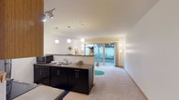 This single-floor space showcases an open-concept layout that seamlessly connects the kitchen and living areas. The kitchen is outfitted with sleek dark cabinetry, a countertop peninsula that doubles as a divider between the cooking and living zones, and a stainless steel sink. A microwave and additional appliances are integrated into the design, while track lighting and a series of globe pendant lights suspended at varying heights provide focused and ambient illumination throughout the kitchen area.

The living space features neutral carpeting and bright white walls, creating a clean and airy atmosphere. A floor lamp anchors one corner, adding a warm glow to complement the abundant natural light that flows in through a large sliding glass door. This door opens directly to an outdoor area surrounded by greenery, blurring the boundary between interior and exterior.

The smooth, white ceiling runs consistently across both areas, enhancing the sense of continuity and openness. The overall ambiance is calm and modern, with a practical layout that balances functional kitchen space with a comfortable, light-filled living area.