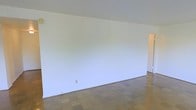 This single-floor space presents a clean, minimalist aesthetic defined by its open layout and bright, airy atmosphere. The main living area is notably spacious, characterized by vaulted ceilings that angle upward to create a sense of added volume and openness. The walls are finished in crisp white throughout, providing a neutral canvas well-suited to a variety of design styles and furnishing arrangements.

The flooring consists of square tile laid in a consistent pattern across the entire visible area, lending a classic, durable quality to the space. The tile's warm, earthy tone offers a subtle contrast to the bright white walls and ceiling, contributing to an overall balanced interior palette.

The room features at least one doorway leading to an adjoining area, suggesting connectivity to additional rooms or spaces within the unit. Electrical outlets are positioned along the base of the walls, and a small wall fixture is visible near the upper corner of the room, indicating functional lighting provisions.

The absence of furnishings in the images allows the architectural qualities of the space — particularly the vaulted ceiling height and generous floor area — to stand out clearly, offering a blank slate with strong foundational design elements.