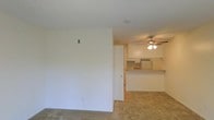 This single-story space features an open-concept layout that connects the living area to the kitchen, creating a cohesive and functional flow throughout. The living area is finished with wall-to-wall carpet in a neutral tone, providing a comfortable and understated foundation for the space. A textured ceiling adds subtle dimension overhead.

The kitchen is partially separated from the living area by a raised breakfast bar with a warm-toned countertop, offering a casual dining or workspace option. Light-colored upper cabinetry extends to the ceiling, maximizing vertical storage. A stainless steel refrigerator is visible within the kitchen, and the space appears to receive adequate ambient lighting.

A ceiling fan with an integrated light fixture is installed in the adjoining area near the kitchen, contributing both air circulation and general illumination. The walls throughout are finished in a clean, neutral white, lending the space a bright and versatile character that would accommodate a variety of furnishing styles.

Overall, the layout is practical and straightforward, suited to everyday living with an emphasis on open sightlines and efficient use of space.