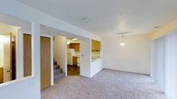 This two-story home offers three bedrooms and two and a half bathrooms across a well-organized floor plan. The main level features an open living area finished with wall-to-wall carpeting, creating a warm and inviting atmosphere. A pass-through opening connects the living space to the kitchen, enhancing the sense of openness while maintaining a defined layout. The kitchen is equipped with warm wood-toned cabinetry, a white refrigerator, and a range, all set atop durable tile flooring that contrasts nicely with the carpeted living area. A pendant light fixture above the pass-through adds a subtle decorative touch. A staircase with carpeted treads leads to the upper level, where the bedrooms and additional bathrooms are located, providing a natural separation between living and sleeping areas. A closet near the staircase offers convenient storage on the main floor. The clean white walls throughout contribute to a bright, neutral aesthetic. With 2.5 bathrooms distributed across both floors, the home supports comfortable day-to-day living for families or shared households.