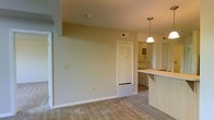 This single-floor residence offers a clean, functional layout with an open-concept design that connects the living area to the kitchen. The living space features wall-to-wall carpeting in a neutral beige tone, light-colored walls, and standard ceiling height, creating a calm and understated ambiance. A floor vent and wall-mounted thermostat control are conveniently positioned, reflecting practical utility throughout.

The kitchen is partially open to the living area via a peninsula-style counter with a light-colored surface, providing both workspace and casual seating potential. A pendant light fixture hangs above, adding a simple decorative touch. The kitchen layout offers good functionality with accessible counter space.

A louvered bifold door leads to an in-unit laundry closet, housing a washer and dryer — a highly convenient feature. Adjacent to this is a doorway providing access to an additional room or hallway, contributing to the overall flow of the unit.

The space maintains a consistent aesthetic throughout, with white trim, neutral wall tones, and standard fixtures. Overall, this residence presents a well-organized, move-in-ready layout suited for comfortable everyday living.