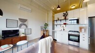 This single-floor urban loft showcases a compelling mix of industrial heritage and contemporary design. The open-plan living and kitchen area sets the tone immediately, with exposed ceiling pipes and conduit painted in a warm gold tone, polished concrete floors that reflect natural light, and a wall-mounted mini-split system providing climate control. The kitchen is efficiently designed with white cabinetry, subway tile backsplash, open wood-and-iron shelving, a gas range, stainless steel dishwasher, and a deep utility sink with a matte black faucet. The living area is anchored by a painted white brick accent wall and expansive black-framed factory windows that flood the space with natural light and offer open city views, lending the room a distinctly loft-like atmosphere. The bedroom continues the industrial aesthetic with an exposed painted brick feature wall, soaring ceiling height, and warm accent lighting, creating an intimate yet airy retreat. A hallway connects the main living spaces to the bathroom, which features a floating wood-toned vanity with an integrated sink, a medicine cabinet mirror, and clean white tile finishes. Polished concrete flooring runs consistently throughout, unifying the space with a cohesive, modern industrial character.