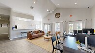 Located at 2074 Midyette Rd in Tallahassee, FL, this single-story community clubhouse offers a well-organized and inviting layout across a compact but efficiently designed floor plan. The space is defined by its striking vaulted ceilings, which soar overhead in the main lounge area and lend the interior a sense of openness and grandeur well beyond its physical footprint. Recessed lighting and a ceiling fan complement the airy feel throughout.

The main lounge area serves as the heart of the space, furnished with a leather sofa, accent chairs, and a jute area rug, creating a comfortable gathering spot. A wall-mounted television and a small beverage station add to its functionality. Adjacent to the lounge is a dedicated office area with multiple workstations and ergonomic seating, ideal for leasing or administrative use.

The kitchen features white shaker-style cabinetry, stone-look countertops with a generous breakfast bar overhang, stainless steel appliances, and black refrigerator. A dining area with a wood table and large multi-pane windows frames picturesque views of a sparkling outdoor pool surrounded by mature Florida greenery, flooding the space with warm natural light. Light wood-look flooring runs throughout, tying all areas together cohesively.