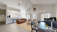 Located at 2074 Midyette Rd in Tallahassee, FL, this single-story community clubhouse offers a well-organized and inviting layout across a compact but efficiently designed floor plan. The space is defined by its striking vaulted ceilings, which soar overhead in the main lounge area and lend the interior a sense of openness and grandeur well beyond its physical footprint. Recessed lighting and a ceiling fan complement the airy feel throughout.

The main lounge area serves as the heart of the space, furnished with a leather sofa, accent chairs, and a jute area rug, creating a comfortable gathering spot. A wall-mounted television and a small beverage station add to its functionality. Adjacent to the lounge is a dedicated office area with multiple workstations and ergonomic seating, ideal for leasing or administrative use.

The kitchen features white shaker-style cabinetry, stone-look countertops with a generous breakfast bar overhang, stainless steel appliances, and black refrigerator. A dining area with a wood table and large multi-pane windows frames picturesque views of a sparkling outdoor pool surrounded by mature Florida greenery, flooding the space with warm natural light. Light wood-look flooring runs throughout, tying all areas together cohesively.