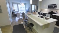 This thoughtfully designed single-floor space showcases a seamless open-concept layout that effortlessly connects the kitchen, dining, and living areas. The kitchen is a standout feature, highlighted by a striking waterfall-edge countertop in a crisp white finish that doubles as a breakfast bar, complemented by dark cabinetry, a modern undermount sink with a contemporary faucet, and clean under-cabinet lighting. Two black iron bar stools add an industrial-chic touch to the bar seating area. The polished concrete floors flow continuously throughout the space, adding an urban, contemporary feel while reflecting the generous natural light that pours in through large windows along the perimeter. The dining area is anchored by a round pedestal table surrounded by wicker-style chairs, topped by a sculptural pendant light that adds warmth and character. Beyond, the living area is comfortably arranged, benefiting from the bright, airy atmosphere created by the oversized windows. The soft grey-blue wall tones provide a neutral, calming backdrop that ties the entire space together. The overall ambiance is modern and inviting, balancing industrial elements with warm, livable touches throughout.