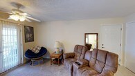 This single-story residence offers a well-organized layout with distinct living, dining, sleeping, and bathing spaces, each thoughtfully finished to create a comfortable and functional environment.

The living room serves as the heart of the home, featuring rich hardwood flooring, a textured ceiling, and a ceiling fan with integrated lighting. Large windows dressed with sheer grommet curtains allow natural light to fill the room, while a wall-mounted flat-screen television and a rustic whitewashed coffee table add character. Dark upholstered sofas provide ample seating, complemented by a distressed wood media console.

The dining area transitions naturally from the living space, showcasing the same hardwood flooring. A round dark-wood table with matching cross-back chairs sits beneath a pendant light, creating an intimate setting for meals.

Two bedrooms feature plush carpeting, ceiling fans, and windows with curtain panels. The primary bedroom includes a whitewashed wood bed frame and matching dresser, while the second bedroom showcases an upholstered headboard and a curated gallery wall arrangement.

Two bathrooms mirror each other in layout, each equipped with a single vanity, built-in mirror, overhead lighting, and a shower-tub combination.