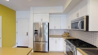 This kitchen is designed with a clean, contemporary aesthetic that combines functionality with modern style. The cabinetry throughout is finished in crisp white with shaker-style doors and brushed hardware, extending from floor to ceiling to maximize storage capacity. A full-height cabinet column frames the stainless steel French door refrigerator with bottom freezer drawer, integrating it seamlessly into the overall design.

The cooking area features a stainless steel gas range with multiple burners, complemented by a built-in stainless steel microwave mounted above the countertops. A subtle patterned tile backsplash in neutral tones runs along the walls between the upper and lower cabinets, adding visual texture without overwhelming the space. The countertops are finished in a warm taupe tone that contrasts tastefully with the white cabinetry below.

The layout is efficient and well-organized, with ample counter space for food preparation. Light-toned flooring contributes to the bright, airy atmosphere of the kitchen. A doorway visible in the background connects the kitchen to adjacent areas of the home, enhancing the sense of flow throughout the single-floor layout.