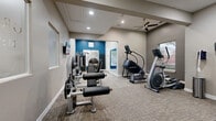 This fitness center offers a thoughtfully designed workout environment across a single floor, providing ample space for a full range of exercise activities. The layout is open and functional, with neutral taupe-toned walls complemented by a striking deep blue accent wall that adds visual interest and energy to the space. Carpet tile flooring runs throughout, providing both comfort and durability underfoot.

The cardio zone is well-appointed with multiple treadmills, a StairMaster, and an elliptical trainer, allowing simultaneous use by several individuals. Strength training is equally represented, with a cable machine system, leg extension and curl machines, a Star Trac Instinct strength unit, free weights, and a flat workout bench. Wall-mounted televisions are positioned throughout the space for entertainment during workouts.

A ceiling fan with recessed lighting provides effective air circulation and illumination, while multiple windows along the perimeter allow natural light to filter in and offer views of the surrounding outdoor environment. Large wall mirrors enhance the sense of space and serve a practical purpose for form monitoring. The overall ambiance is clean, modern, and purposefully arranged to support a comprehensive fitness routine.