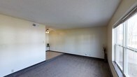 This single-floor space presents a straightforward, functional layout with a notably open living area. The room features a combination of grey carpeting and hard flooring, delineated by a subtle transition that helps define separate zones within the open floor plan. Neutral, light-toned walls create a clean and versatile backdrop, offering flexibility for a variety of interior styles.

A ceiling fan is centrally positioned, providing both lighting and air circulation to the main living area. The textured popcorn ceiling adds a classic touch, while dark baseboards provide a modest contrast to the pale walls. A wall-mounted air vent is visible, indicating a central HVAC system.

The space benefits from a simple, uncluttered design that maximizes usable floor area. The open layout allows for flexible furniture arrangements, making it adaptable for various needs. Overall, the space offers a practical and livable environment with neutral finishes that serve as a blank canvas for personalization.