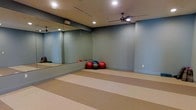 This single-floor fitness studio is thoughtfully designed to support a variety of exercise and wellness activities. The open layout provides ample room for movement, with a large full-wall mirror along one side that enhances the sense of space while allowing practitioners to monitor their form during workouts. The walls are finished in a calming muted blue-gray tone, creating a focused yet relaxed atmosphere well-suited for yoga, pilates, stretching, or group fitness classes. The flooring features a two-tone striped design in neutral beige tones, offering a clean and practical surface for floor exercises. Recessed lighting is evenly distributed across the ceiling, ensuring the space is well-lit throughout, while ceiling fans provide consistent air circulation to keep the environment comfortable during active sessions. A collection of exercise balls in various sizes is stored neatly in the corner, indicating the studio's readiness for a range of fitness activities. The overall design is minimal and functional, free of clutter, with clean lines and a practical layout that prioritizes utility and ease of use for fitness enthusiasts of all levels.
