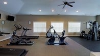 This dedicated fitness center is thoughtfully designed to provide a comfortable and functional workout environment. The space is outfitted with a selection of cardio equipment, including a treadmill, elliptical trainer, and stationary recumbent bike, all positioned to allow for easy use and movement between machines. Protective rubber floor mats are placed beneath each piece of equipment, complementing the durable, textured flooring throughout the room.

Natural light fills the space through a row of large windows fitted with white horizontal blinds, creating a bright and energizing atmosphere while offering control over light levels. Recessed ceiling lights provide additional illumination, ensuring the space remains well-lit at all times. A ceiling fan with a classic multi-blade design circulates air efficiently, keeping the environment cool and comfortable during workouts.

The walls are finished in a neutral warm tone, contributing to a clean and welcoming aesthetic. An informational posting on the wall near the entrance provides facility guidelines for users. The overall layout offers generous open floor space, allowing room for stretching or additional freestanding equipment. This fitness center is a practical and well-appointed amenity suited for a consistent and well-rounded exercise routine.
